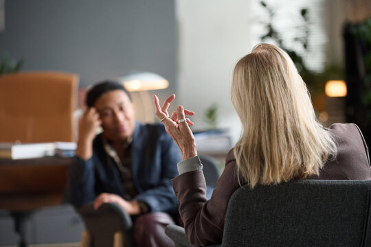Senior Caucasian woman gesturing while conducting therapy session with young Black woman listening attentively in office setting, both seated and engaged in conversation