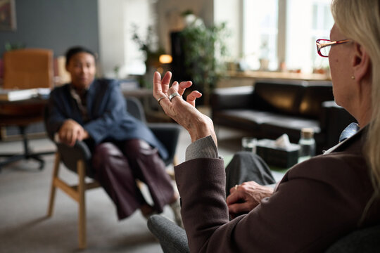 Elderly Caucasian woman gesturing while conducting therapy session with young Black woman sitting and listening in modern office setting, focus on therapist hand movement