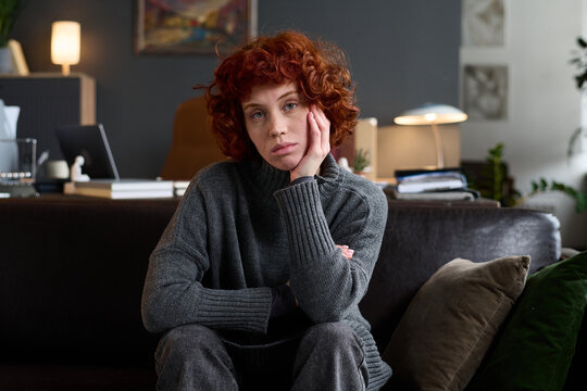 Portrait of young adult Caucasian woman sitting on sofa resting head on hand looking at camera during therapy session in modern office setting with thoughtful expression