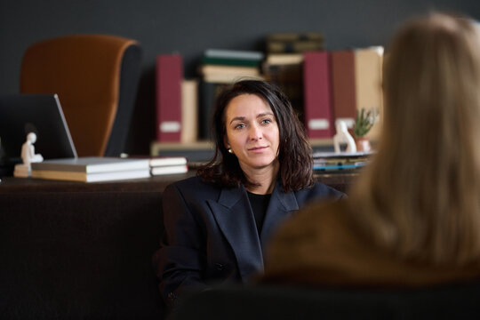 Portrait of Caucasian woman sitting in office setting engaging in therapy session with another woman visible from behind, bookshelf and desk in background