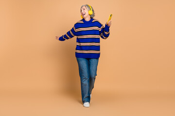 Joyful mature woman dancing with a yellow phone and headphones in a striped knit sweater against a...