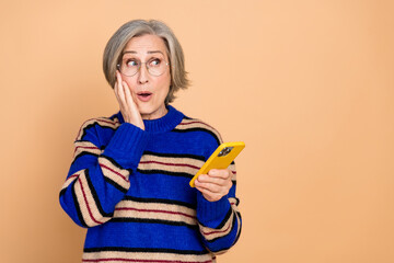 Aged mature woman wearing a blue striped sweater surprised while looking at her yellow smartphone...