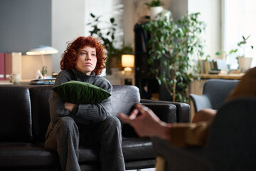 Caucasian young adult woman sitting on sofa hugging pillow during therapy session, listening to therapist whose hands are visible in foreground, indoor counseling setting