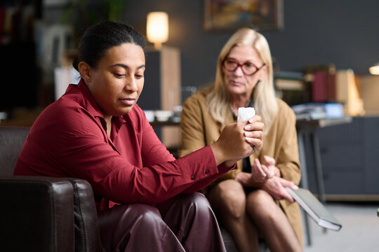 Young Black woman sitting on couch holding tissue, senior Caucasian therapist listening attentively in office during therapy session, bookshelf and lamp in background