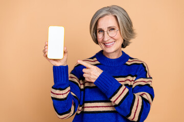 Mature woman with grey hair and glasses holds a blank smartphone and smiles while pointing at the...