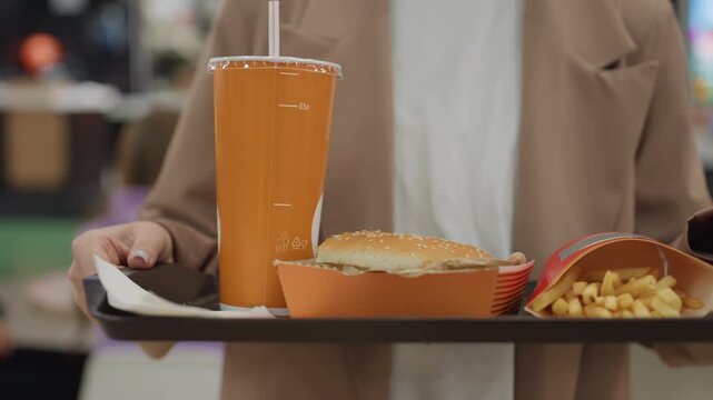 caucasian woman carrying fast food tray with burger fries and drink in busy mall food court, beige coat, closeup hands and manicured nails, neutral commuter scene with background bustle and counter