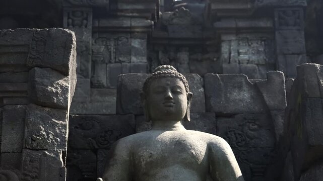 Statues of Buddha sitting in Vara mudra inside wall niches illuminated by sun. Beautiful decorative details of Borobudur Mahayana Buddhist temple, Magelang, Central Java, Indonesia. Camera zooms out.