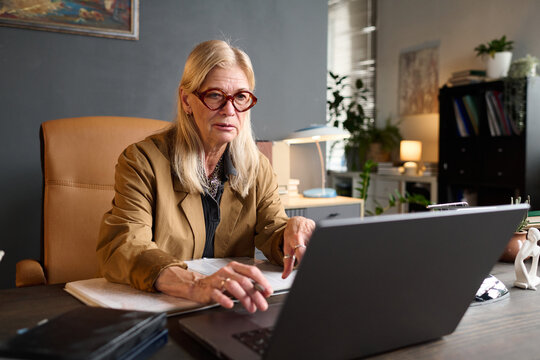 Senior Caucasian woman sitting at desk working on laptop, wearing glasses, appearing focused while conducting therapy session online, office background with documents and books visible