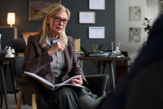 Caucasian senior woman therapist conducting counseling session holding notebook gesturing while listening to client in office setting providing professional mental health support