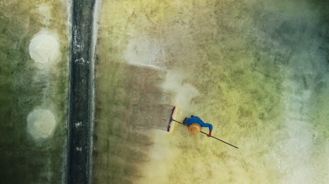 Top down drone view of Asian worker using rake to move salt crystals in shallow evaporation pond. Abstract texture of traditional sea salt production with copy space.