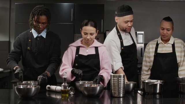 Female culinary course trainee mixing ingredients in bowl next to Black male groupmate while chef instructing other students during class in restaurant kitchen