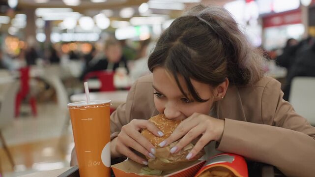 teenage girl enthusiastically consumes hefty burger, young girl excitedly bites into huge sandwich at food court, adolescence girl eagerly indulges in substantial burger with vibrant atmosphere