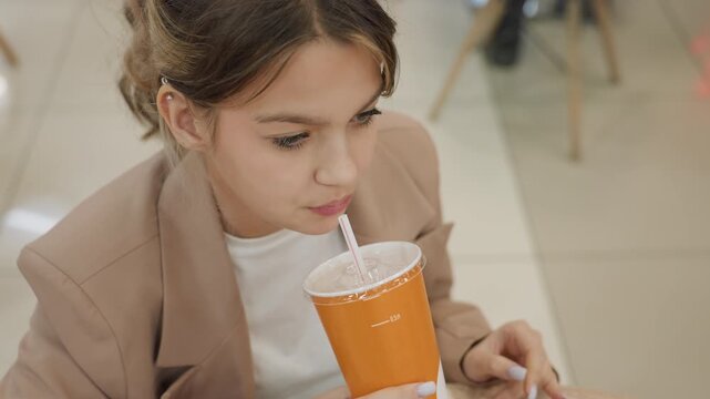 teen caucasian student sipping drink at mall table, orange cup with straw, blazer outfit, thoughtful expression, casual hands on cup, indoor food court background, soft daylight, quiet pause