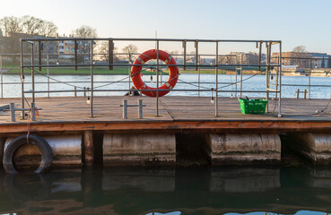 Fototapeta premium Floating pier with metal railings, a bright lifebuoy ring, and mooring hardware above still water. Simple geometric composition for safety, rescue, waterfront infrastructure, and maritime travel conce