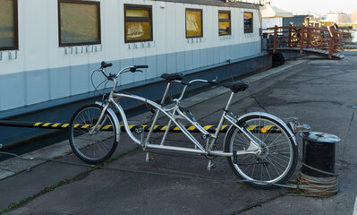 Fototapeta premium Modern silver tandem bicycle standing on a dockside pavement beside moored boats. Clean lines and travel vibe suggest leisure, cycling, and sustainable transport near water, perfect for mobility and o