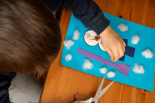 Overhead view of a child placing paper elements on a snowman collage. Adjusting the purple broom next to the button body