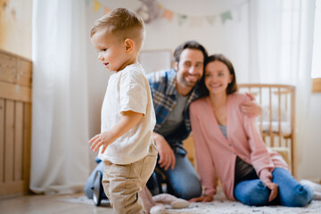Happy young caucasian parents mother and father watching how their small little kid child toddler infant baby making first steps at home, learning how to walk. Parenthood and childcare