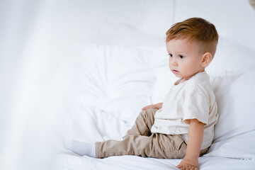 Cute little small child caucasian infant toddler newborn boy son sitting relaxing on the white bed. Teething concept, childcare parenthood, copy space. Advertisement for children's products, clothing