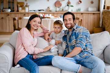 Happy caucasian cheerful young family of three looking at camera spending time together at home. Parents and toddler at home on the couch. Parenthood concept. Insurance mortgage for people