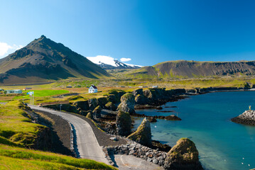 A winding road follows Iceland’s rugged coastline toward a small white church, with green hills,...
