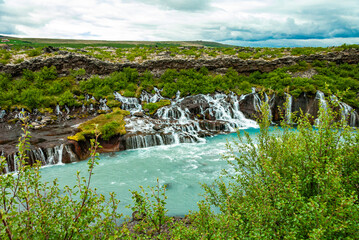 Hraunfossar is a unique Icelandic waterfall in West Iceland where clear, cold spring water seeps directly from under the Hallmundarhraun lava field, forming hundreds of small, delicate waterfalls.