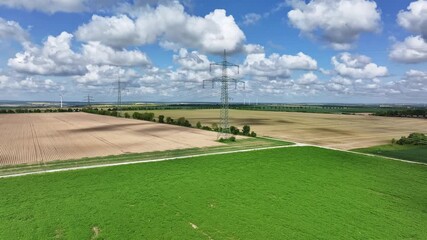 Slow side drone flight around a high-voltage pylon with the transmission line crossing the landscape, surrounded by cultivated and harvested fields under a blue sky with clouds.