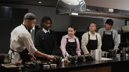Multiethnic group of trainees wearing black aprons learning new recipe in culinary course class led by professional chef demonstrating ingredients on workbench in restaurant kitchen