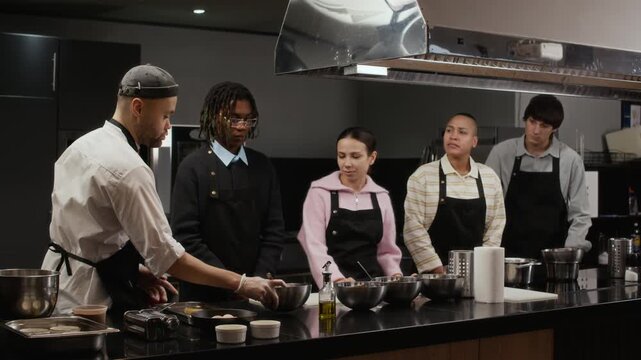 Multiethnic group of trainees wearing black aprons learning new recipe in culinary course class led by professional chef demonstrating ingredients on workbench in restaurant kitchen