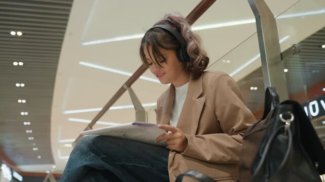 young woman headphones studying notebook on mezzanine bench under warm led lights, leather bag beside, focused expression, slow camera pan captures concentration and urban architecture