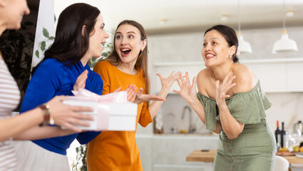 Happy surprised young Asian woman receiving unexpected gift from besties coming to celebrate birthday bash in cozy home setting with set table in background..