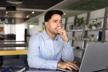 Serious thoughtful Latin entrepreneur man working on online business project in co-working space, typing on laptop, touching chin, looking away in deep thoughts, thinking on marketing strategy