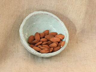 Handmade ceramic bowl filled with raw almonds, photographed from above on natural linen fabric. Minimalist food still life with organic textures, calm mood and healthy lifestyle concept.