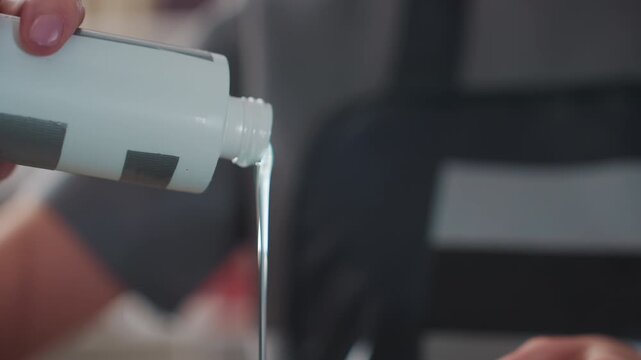 assistant pouring precise dose from bottle, steady stream measured over mixing bowl, apron and tools visible in background, closeup of careful hand and flowing liquid, routine salon calibration