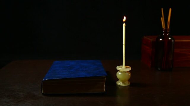 Burning wax white candle with spell book on the wood table, wooden box and bag on dark black background with empty copy space. Ancient old magic objects