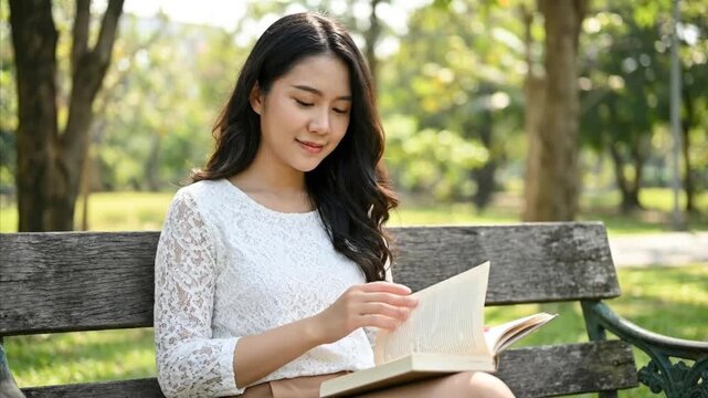 A beautiful young woman with long dark hair is captured in a serene outdoor setting, gracefully turning the pages of a book while sitting on a rustic wooden park bench. Bathed in soft daylight, she ap