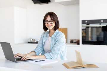 A young woman with glasses is sitting at a kitchen table, working on her laptop. She looks happy and relaxed while a book lies open nearby.