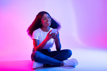 A young black lady sits cross-legged on the floor, smiling as she browses social media on her smartphone in a lively neon light environment. She is engaged in messaging a friend.