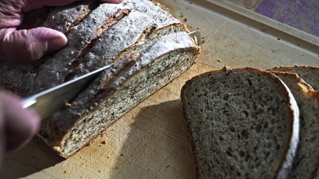 A person cuts slices from a loaf of homemade bread on a wooden cutting board. The sunlight shines on the bread as each slice is carefully removed, creating perfect pieces for sandwiches.