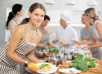Young girl serving cooked chicken breast with mustard standing surrounded by other female members during cooking master class