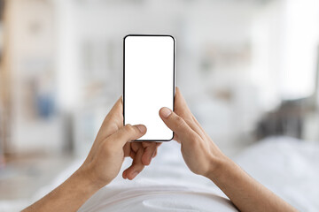 A man lies comfortably in bed, holding a blank smartphone with a white screen. He appears relaxed and enjoys his cozy space. This scene captures a peaceful moment at home.