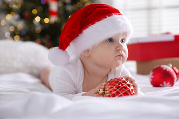 Cute little baby in Santa hat and baubles on bed at home. Christmas season
