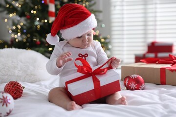 Cute little baby in Santa hat with gift box and baubles on bed at home. Christmas season