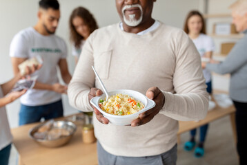 An African American senior man holds a plate filled with a warm meal inside a charity office. He is receiving food from volunteers dedicated to supporting elderly and homeless communities.