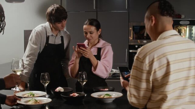 Smiling young woman taking selfie with groupmate while celebrating successful culinary class with white wine and delicious dishes in professional kitchen