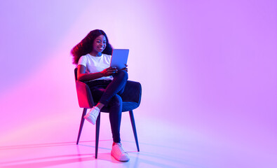 A cheerful young black lady sits comfortably in an armchair, using a tablet to study or work remotely. The vibrant neon light creates a lively atmosphere, enhancing her focus.