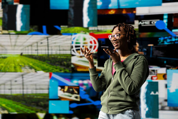 Fototapeta premium Happy woman records voice note on smartphone, discussing movies with friend, standing in front of vibrant video wall collage. Woman excited to share audio message to friend on phone