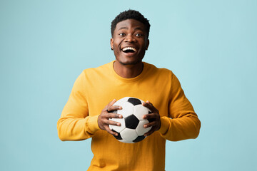 Happy millennial black guy in casual with soccer ball posing on blue studio background, emotional african american young man football fan going to game, copy space. Sport concept © Prostock-studio