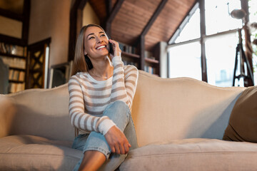 Fototapeta premium A young woman smiles broadly as she talks on her phone, relaxed on a comfortable couch in a bright and inviting living room filled with natural light.