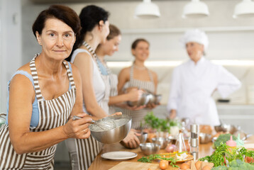 Aged female participiant of cooking master class holding bowl and whisk in her hands standing around other female members