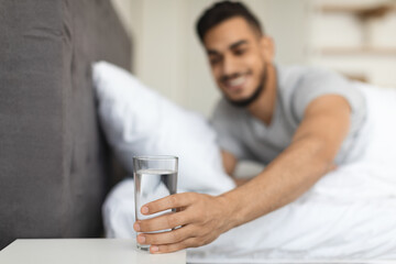 A young Middle Eastern man smiles as he reaches for a glass of water on the bedside table in his bedroom. It is morning and he has just woken up feeling thirsty and refreshed.